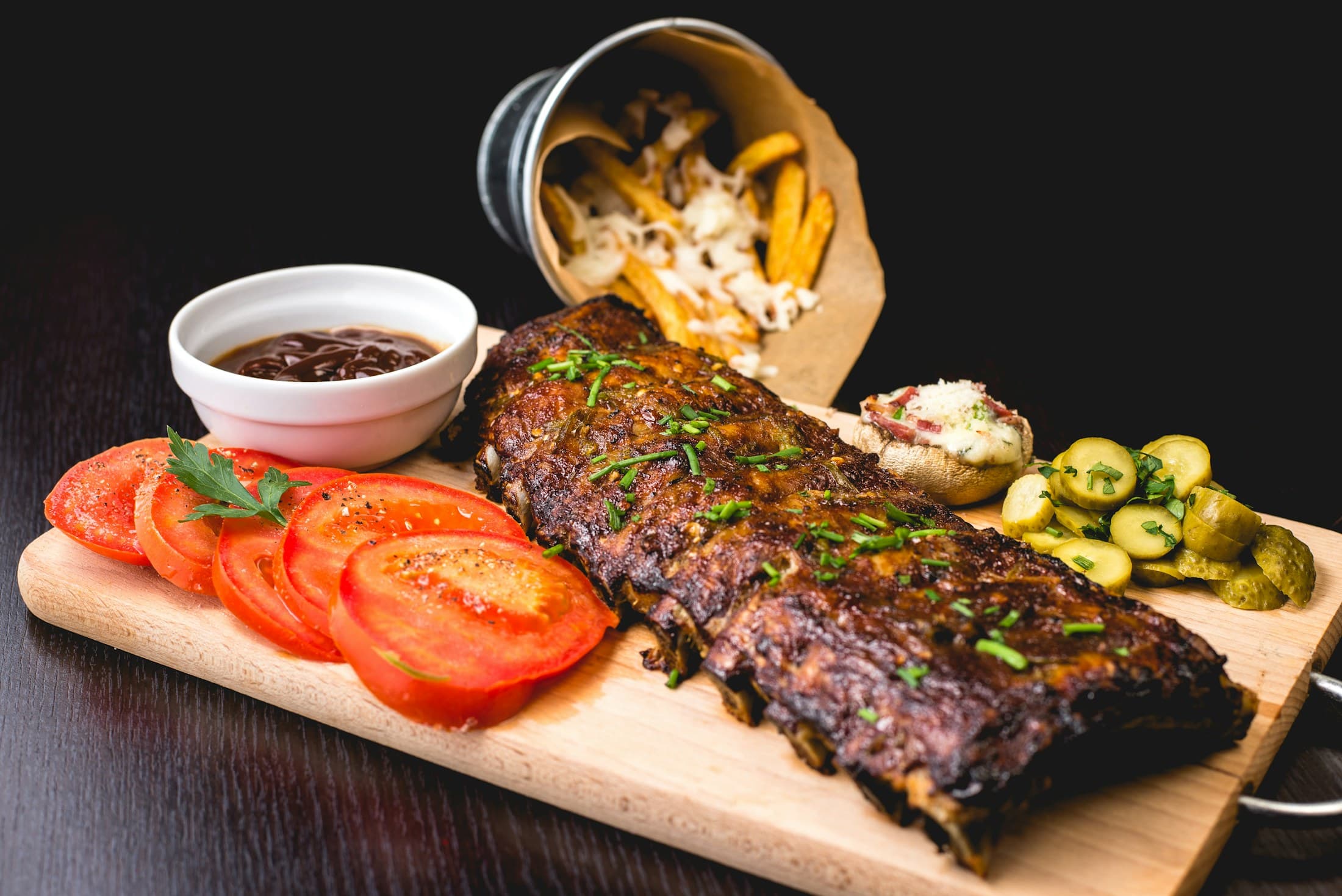 A rack of glazed ribs with tomato slices and a small bowl of sauce.