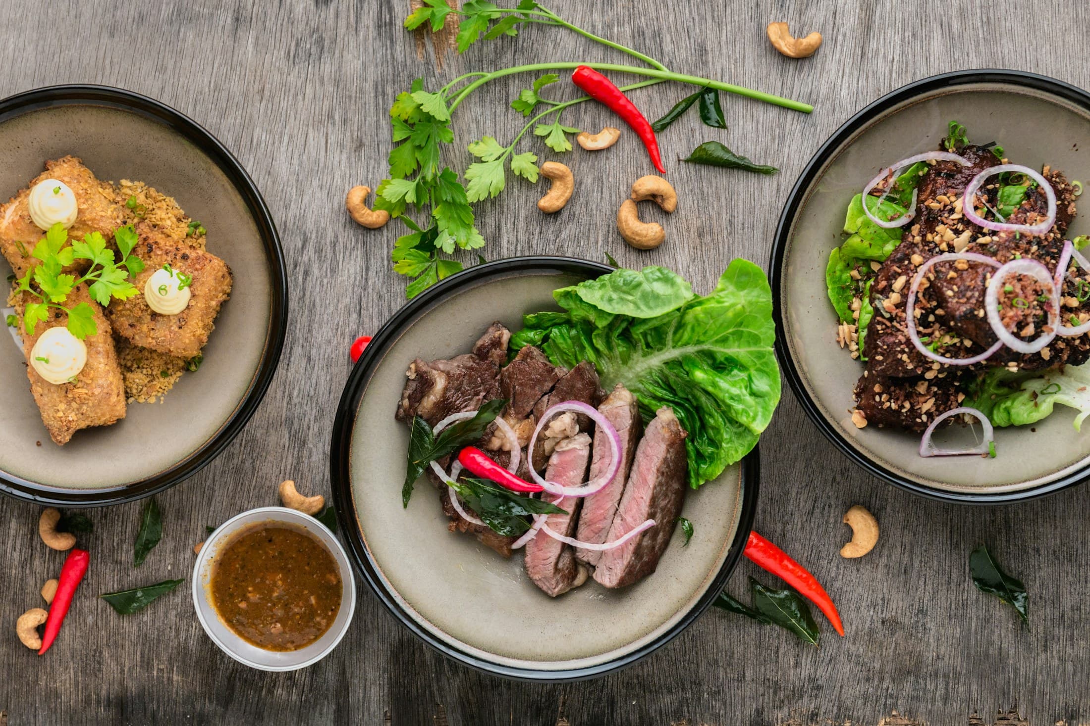 Plates of sliced grilled beef with greens, chillies and sauces, seen from above.