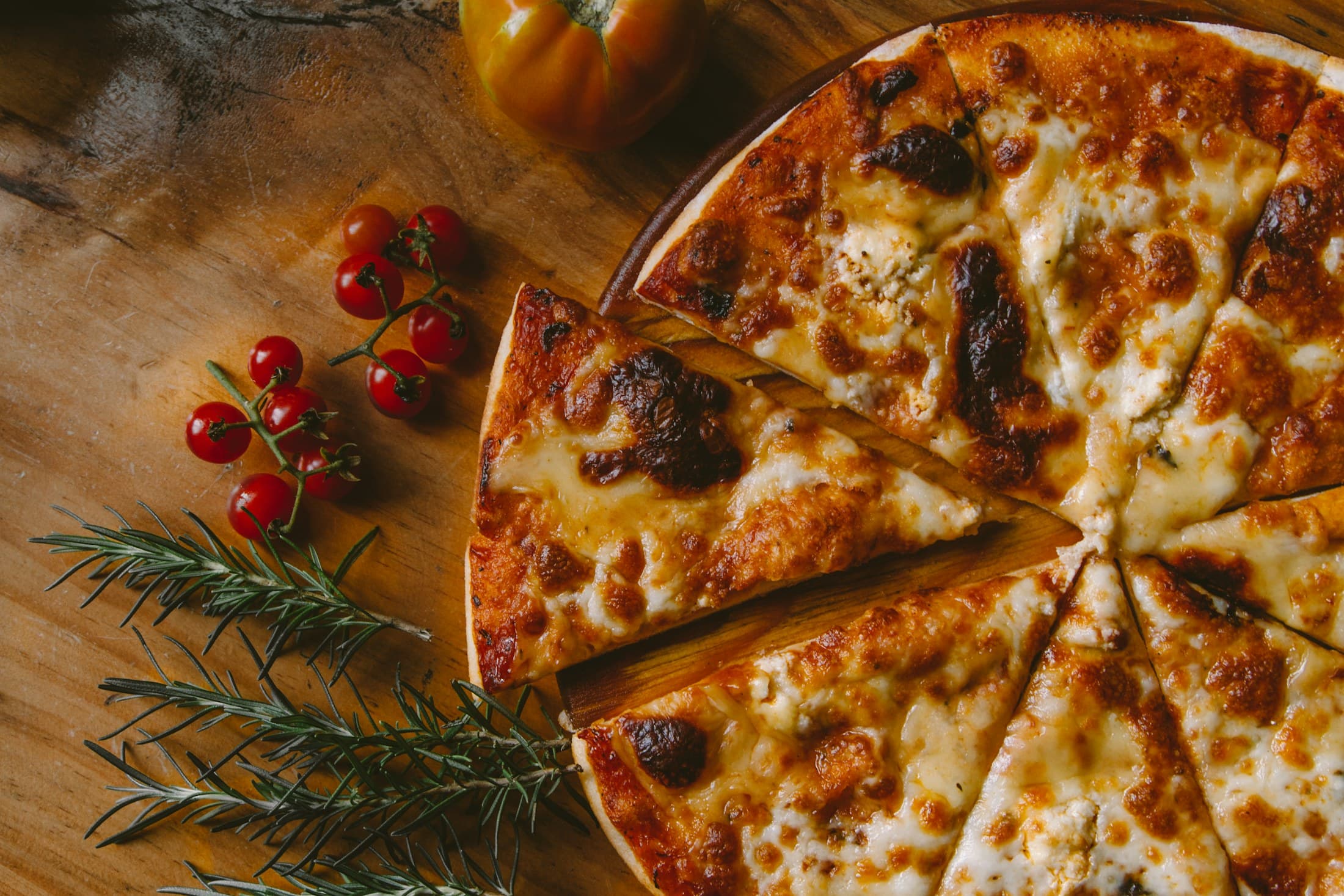 A wood-fired pizza with blistered crust beside cherry tomatoes and rosemary.